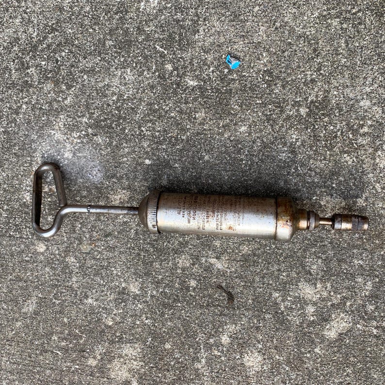 May include: A vintage grease gun with a silver metal body and a handle. The gun has a nozzle on one end and a handle on the other. The body of the gun has text printed on it. The grease gun is designed for applying grease to machinery.