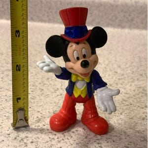 Pu&ograve; includere: Una statuetta di Topolino, alta circa 7,6 cm, con un cappello a cilindro rosso con dettagli blu, una giacca blu, pantaloni rossi e un gilet giallo. La statuetta ha guanti bianchi e scarpe rosse. La statuetta &egrave; in posa con un braccio teso.