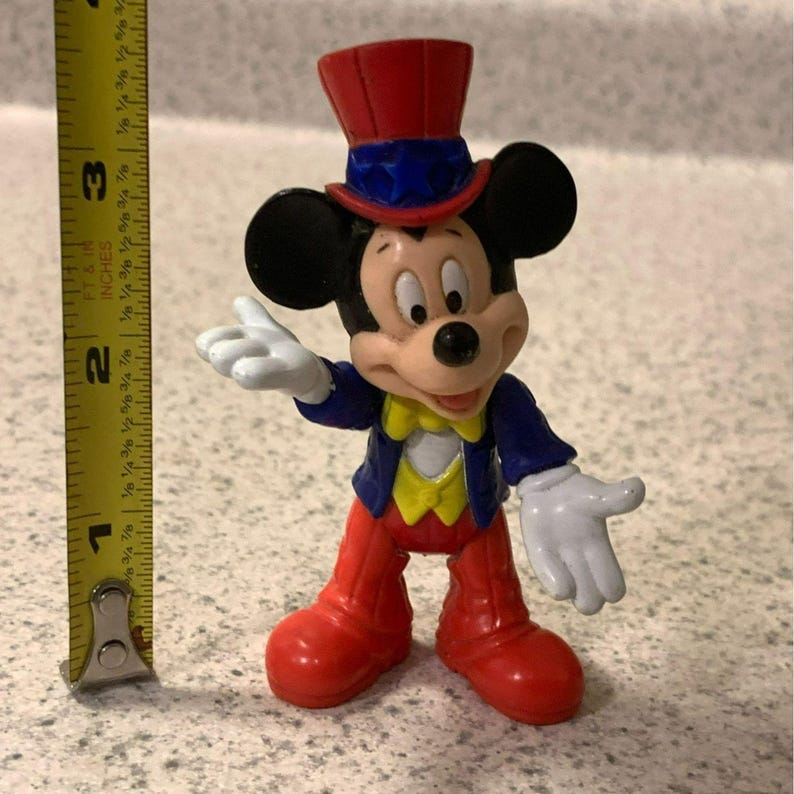 Pu&ograve; includere: Una statuetta di Topolino, alta circa 7,6 cm, con un cappello a cilindro rosso con dettagli blu, una giacca blu, pantaloni rossi e un gilet giallo. La statuetta ha guanti bianchi e scarpe rosse. La statuetta &egrave; in posa con un braccio teso.