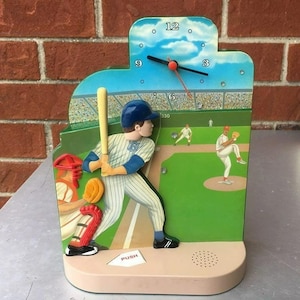 May include: A baseball-themed alarm clock with a green and brown base. The clock face is blue with white numbers and a red second hand. The clock features a baseball player at bat, a catcher, and a pitcher. The clock has a push button on the base and a speaker.