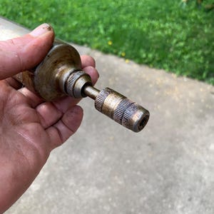 May include: A vintage, cylindrical metal tool with a textured grip and a smaller, ribbed end. The tool has a worn, aged appearance, suggesting it has been used. The metal has a tarnished, bronze-like color.