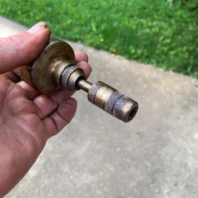 May include: A vintage, cylindrical metal tool with a textured grip and a smaller, ribbed end. The tool has a worn, aged appearance, suggesting it has been used. The metal has a tarnished, bronze-like color.