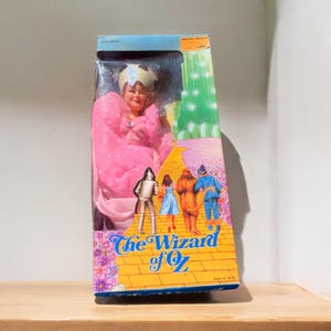 May include: A vintage doll of a character from "The Wizard of Oz" in its original box. The doll is wearing a pink dress and is in a clear plastic window. The box features illustrations of the yellow brick road and characters from the movie. The text "The Wizard of Oz" is displayed on the box.