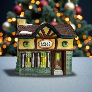 May include: A miniature ceramic clock maker's shop with a brown roof, yellow walls, and green accents. The shop features a sign that reads "CLOCK MAKER" and is decorated with wreaths. The background is a blurred Christmas tree.