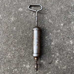 May include: An antique, cylindrical grease gun made of metal, featuring a handle and nozzle. The body of the gun shows signs of wear and tear, with visible text. The overall color is a mix of silver and brown, suggesting age and use.