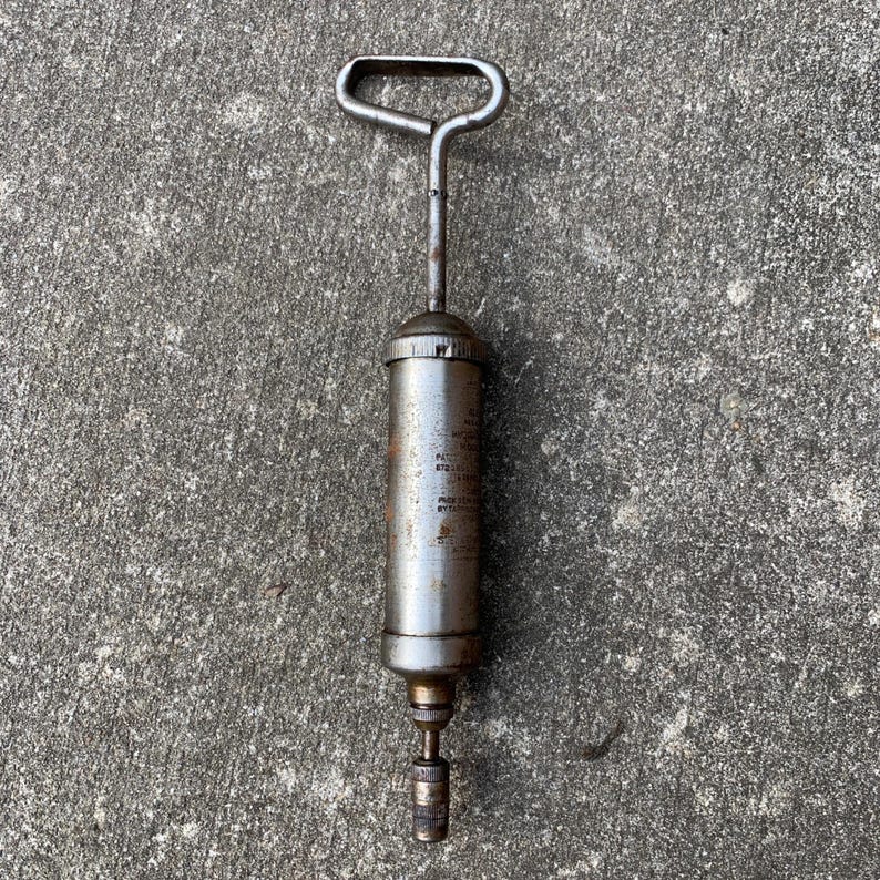 May include: An antique, cylindrical grease gun made of metal, featuring a handle and nozzle. The body of the gun shows signs of wear and tear, with visible text. The overall color is a mix of silver and brown, suggesting age and use.