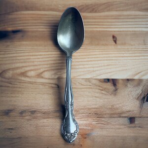 May include: A silver spoon with an ornate handle, resting on a wooden surface.