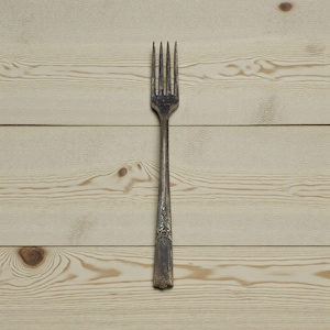May include: A vintage silver fork with four tines, resting on a light-colored wooden surface. The fork has an ornate design on the handle and shows signs of wear, suggesting age and use. The wood grain is visible.