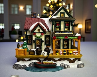 May include: A detailed Christmas village figurine depicting a marine supply shop. The shop has a red roof with snow, green trim, and a sign that reads "MARINE SUPPLIES". A small boat sits in the water in front of the shop.