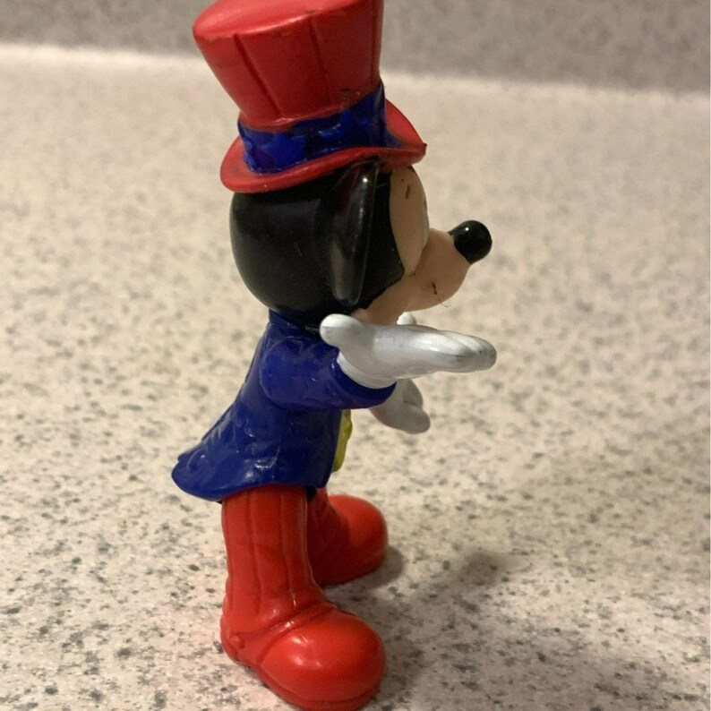 Pu&ograve; includere: Una piccola statuina di un topo dei cartoni animati con un cappello a cilindro rosso con una fascia blu, una giacca blu, stivali rossi e guanti bianchi. Il topo ha orecchie nere e un naso nero. La statuina &egrave; su una superficie chiara.