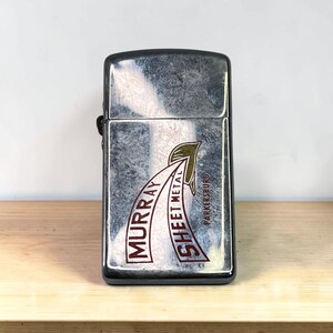 May include: A rectangular silver-toned metal lighter with a hinged lid. The front surface features a graphic design with a green leaf icon and the printed text MURRAY SHEET METAL PARKERSBURG in dark red lettering against a brushed metal finish.