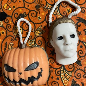 May include: Two Halloween-themed ornaments, a pumpkin with a carved face and a white mask with a scary expression. Both ornaments are hanging from white rope.