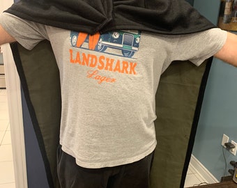 May include: A person wearing a black and green hooded cape over a grey t-shirt with a vintage car graphic and the text "LANDSHARK Lager".