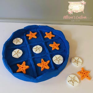 May include: A handmade blue Tic-Tac-Toe game with orange starfish and white sand dollar game pieces. The game is on a white surface. The game pieces are scattered around the board.