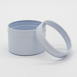 May include: White metal tin with a clear lid. The tin is round and has a smooth surface. The lid is slightly domed and has a clear plastic window.