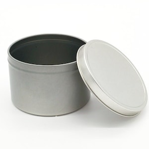 May include: A silver metal tin with a lid. The tin is round and has a smooth surface. The lid is slightly domed and has a rolled edge.