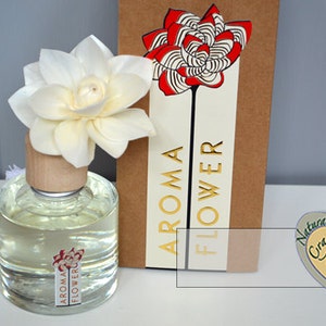 May include: Aroma flower diffuser with a white flower top and a clear glass bottle. The bottle has a wooden cap and a label with the words "Aroma Flower". A brown card with a red and white flower design is behind the bottle.