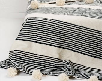 Puede incluir: Una manta de lana a rayas blancas y negras con pompones blancos en los bordes. La manta está doblada en una cama con dos almohadas blancas con pompones.