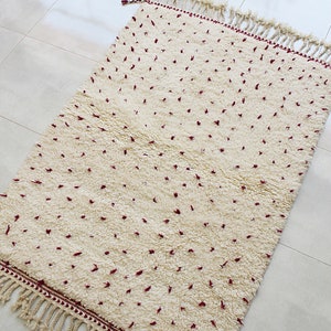 May include: Cream-colored rectangular rug with a plush texture and a pattern of small, scattered red dots. The rug has a fringe border and is suitable for home decor.