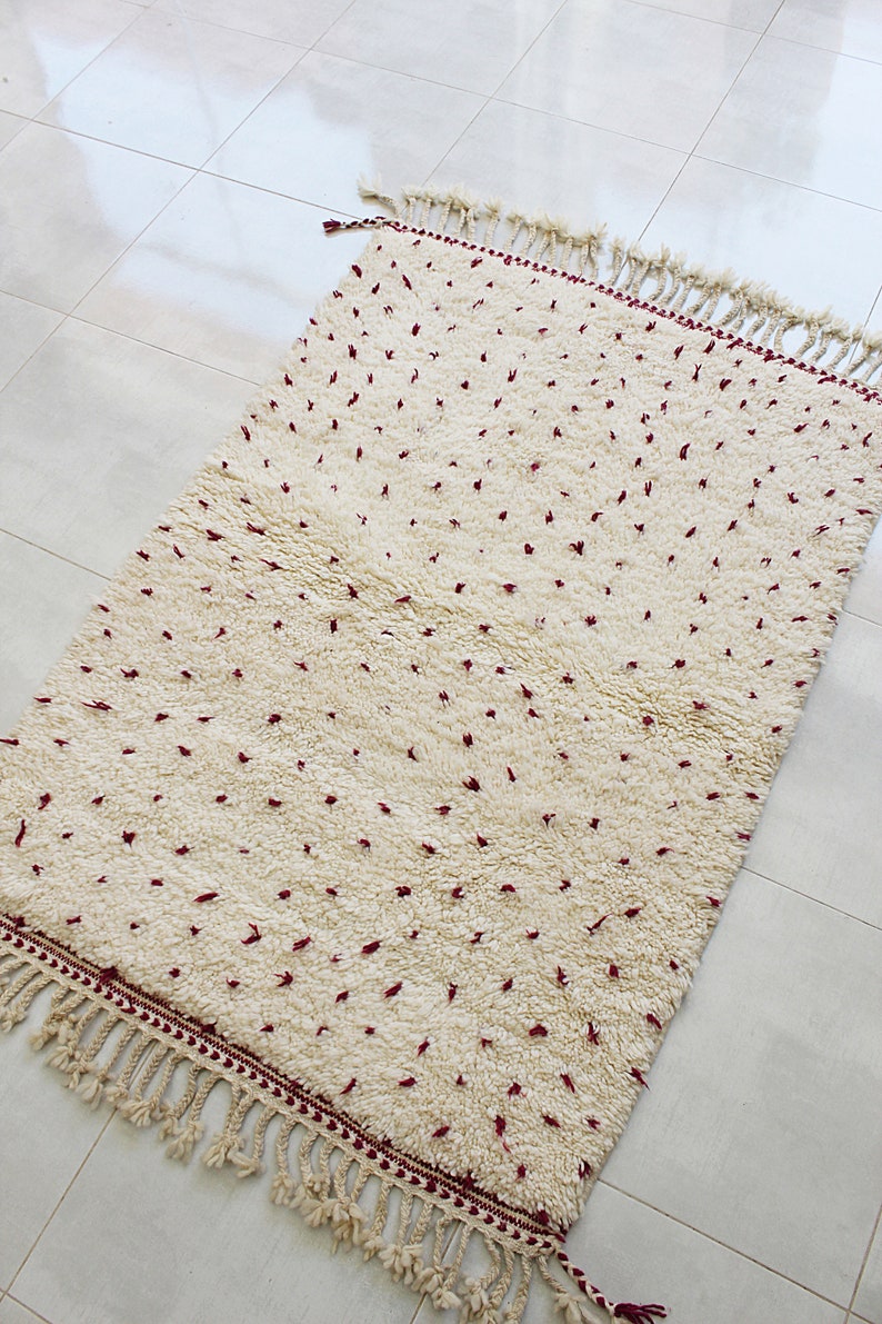 May include: Cream-colored rectangular rug with a plush texture and a pattern of small, scattered red dots. The rug has a fringe border and is suitable for home decor.