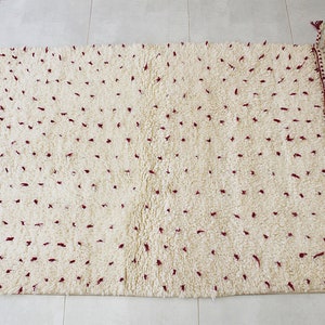 May include: Cream-colored, rectangular rug with a plush, textured surface and scattered maroon accents. The edges are finished with a decorative border and fringe detailing, adding a touch of elegance to this floor covering.