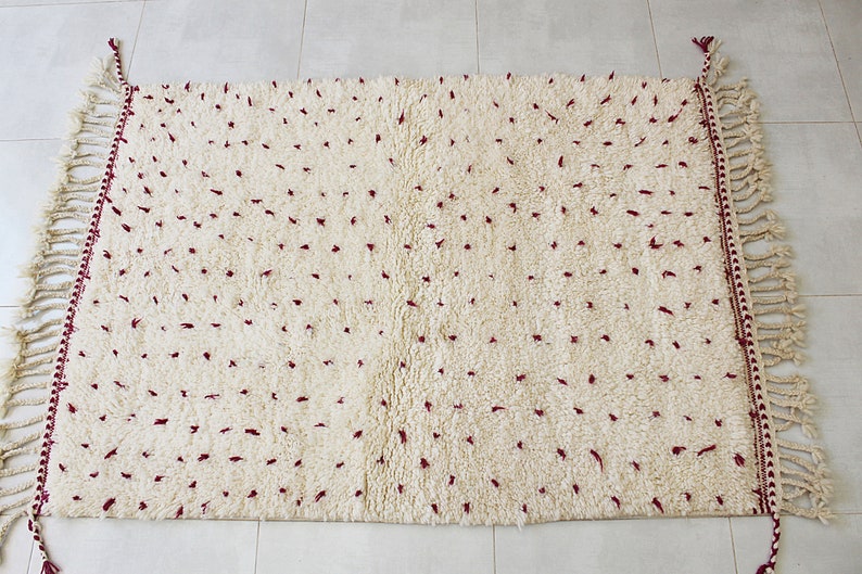 May include: Cream-colored, rectangular rug with a plush, textured surface and scattered maroon accents. The edges are finished with a decorative border and fringe detailing, adding a touch of elegance to this floor covering.