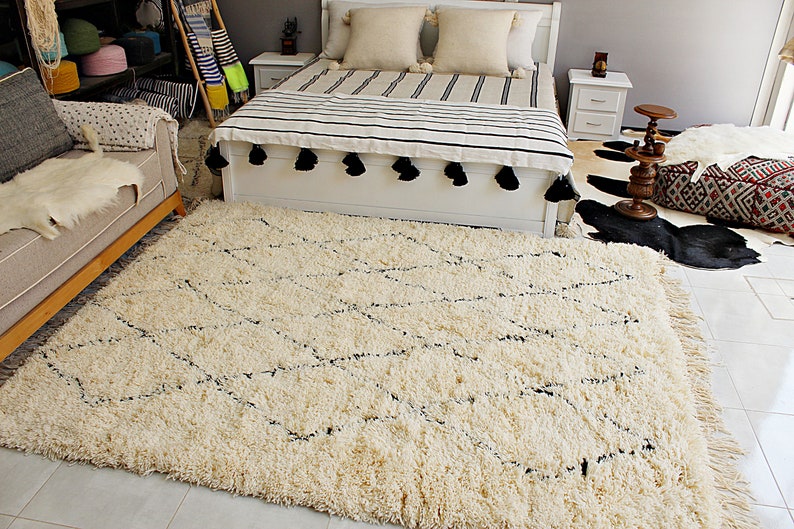 May include: A white shag rug with a black diamond pattern. The rug is in a room with a white bed, a black and white cowhide rug, and a brown wooden stool.