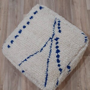 K&ouml;nnte beinhalten: Ein quadratischer Pouf in Wei&szlig; und Blau mit geometrischem Muster. Der Pouf ist aus flauschigem, weichem Material gefertigt und hat einen blauen Rand.