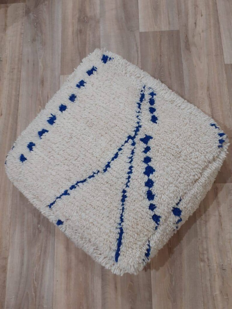 K&ouml;nnte beinhalten: Ein quadratischer Pouf in Wei&szlig; und Blau mit geometrischem Muster. Der Pouf ist aus flauschigem, weichem Material gefertigt und hat einen blauen Rand.