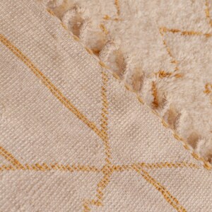 May include: Close-up of a cream-colored woven rug with a subtle geometric pattern in a light yellow color. The rug has a fringe border.