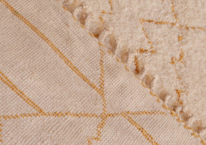 May include: Close-up of a cream-colored woven rug with a subtle geometric pattern in a light yellow color. The rug has a fringe border.