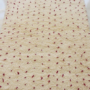 May include: Cream-colored, rectangular rug with a textured, plush surface. The rug features small, scattered burgundy accents and a burgundy border with fringe along the bottom edge. A decorative floor covering.