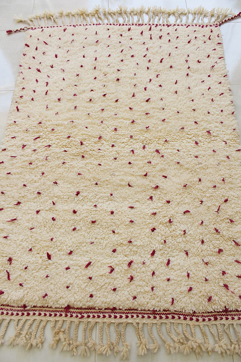May include: Cream-colored, rectangular rug with a textured, plush surface. The rug features small, scattered burgundy accents and a burgundy border with fringe along the bottom edge. A decorative floor covering.