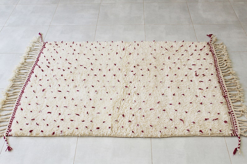 May include: Cream-colored, rectangular rug with a plush texture and scattered burgundy accents. The rug features fringed edges and burgundy trim along the sides. A decorative floor covering for a living room or bedroom.