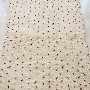 May include: Cream-colored rug with a textured, dotted pattern in burgundy. The rug has a fringe border and tassels at the ends. This decorative rug is suitable for living rooms, bedrooms, or entryways.