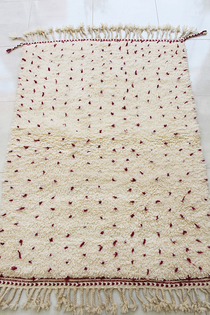 May include: Cream-colored rug with a textured, dotted pattern in burgundy. The rug has a fringe border and tassels at the ends. This decorative rug is suitable for living rooms, bedrooms, or entryways.
