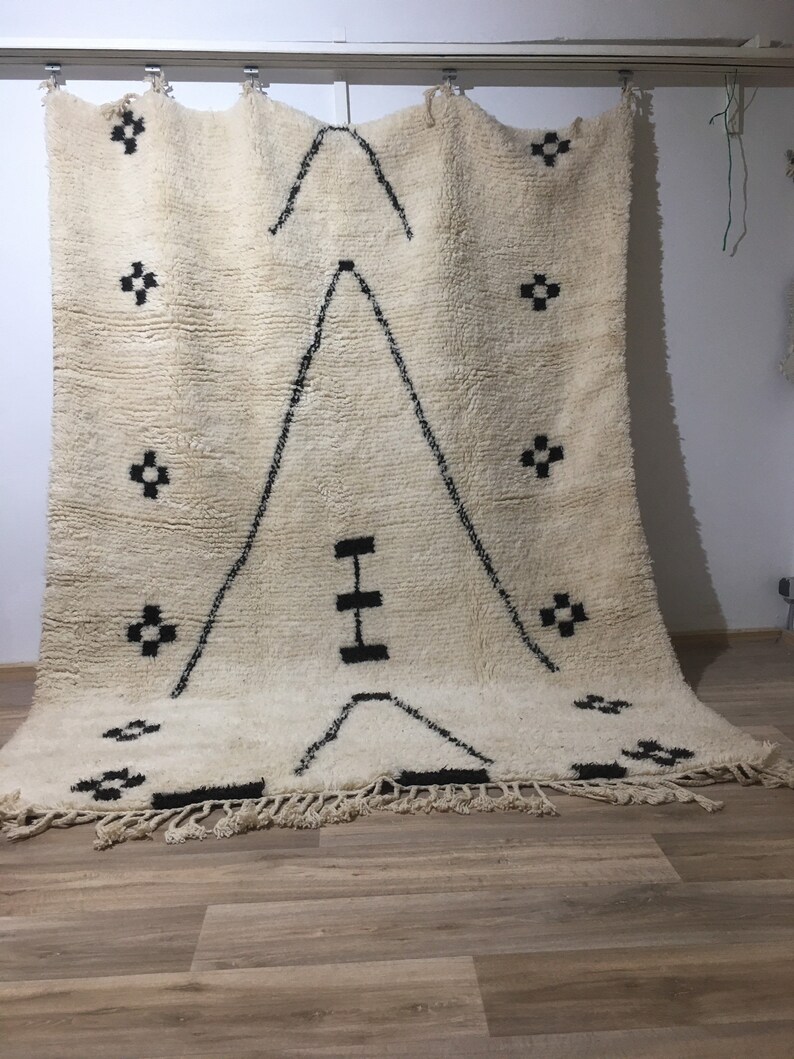 Puede incluir: Una alfombra marroqu&iacute; blanca con un patr&oacute;n geom&eacute;trico negro. La alfombra tiene un borde de flecos y est&aacute; colgada en una pared.