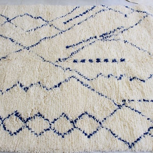 Könnte beinhalten: Ein dicker, cremefarbener Hochflorteppich mit handgeknüpfter Textur und geometrischen Mustern in Dunkelblau. Das Design umfasst miteinander verbundene Rautenformen, gezackte Linien und einen vertikalen blauen Rand an der rechten Kante.