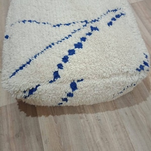 K&ouml;nnte beinhalten: Wei&szlig;er Bodenkissen mit einem blauen geometrischen Muster. Das Kissen ist aus einem weichen, flauschigen Material.