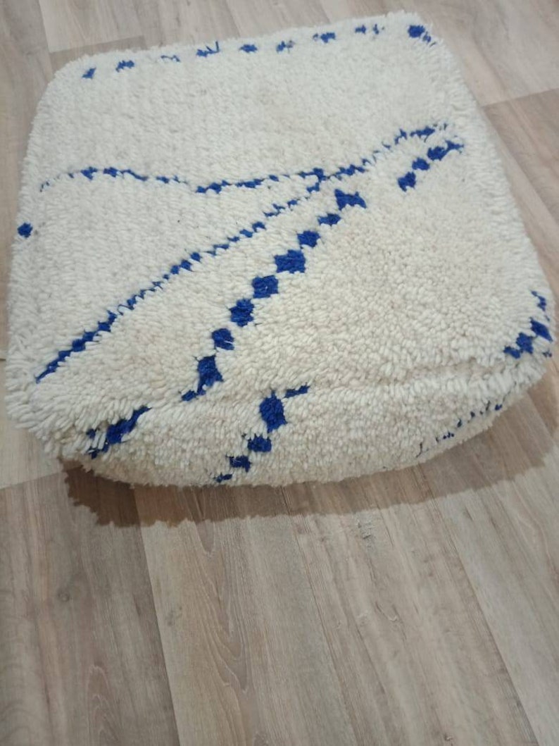 K&ouml;nnte beinhalten: Wei&szlig;er Bodenkissen mit einem blauen geometrischen Muster. Das Kissen ist aus einem weichen, flauschigen Material.