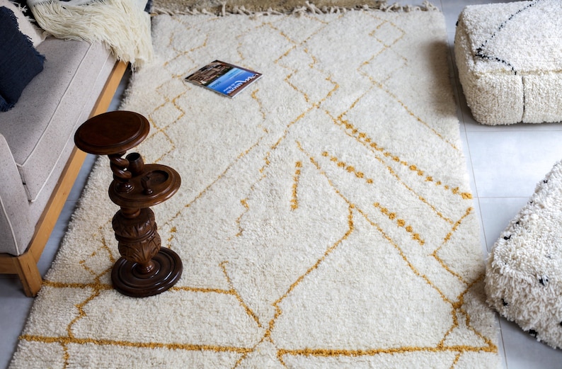 May include: A white shag rug with a geometric pattern in a light gold color. The rug is in a living room setting with a couch, a wooden side table, and two poufs.