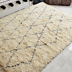 May include: A cream-colored rug with a black diamond pattern. The rug has a shaggy texture and is made of wool.