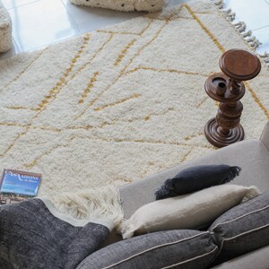 May include: A white and gold patterned rug with a wooden stand and a book titled "Maisons du Maroc" on a couch with black and white pillows.