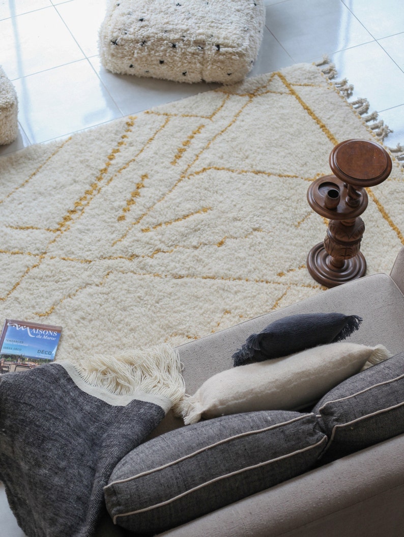 May include: A white and gold patterned rug with a wooden stand and a book titled "Maisons du Maroc" on a couch with black and white pillows.