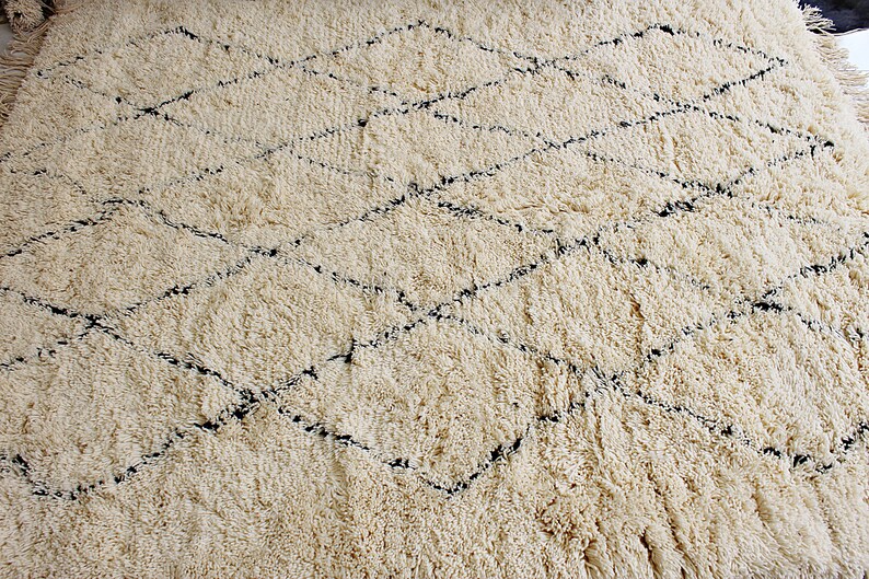 May include: A cream-colored rug with a diamond pattern woven in black thread. The rug has a fluffy, textured surface.