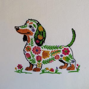 May include: Embroidered dachshund dog design with floral patterns in various colors. The dog's body is white, with brown paws and a black tail. The floral patterns include pink, orange, and green. The dog is standing on a patch of green grass with small pink flowers.