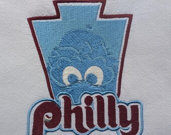May include: Embroidered patch featuring a blue, cartoon-like creature with large white eyes, set within a light blue and maroon outline. The word "Philly" is embroidered in maroon with a light blue border, all on a white fabric.