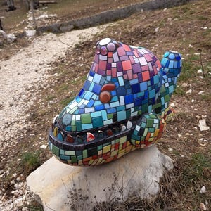 May include: A colorful mosaic sculpture of a creature with a large head, teeth, and a tail. The sculpture is made of small, square tiles in shades of blue, green, red, and purple. It sits on a large, gray rock.