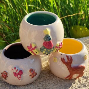 Peut inclure: Trois pots de fleurs en céramique sur le thème de Noël avec un glaçage blanc. Le pot de fleurs de gauche a un motif de canne en sucre et d'homme en pain d'épices. Le pot de fleurs du milieu a un motif de sapin de Noël avec des cadeaux. Le pot de fleurs de droite a un motif de renne.