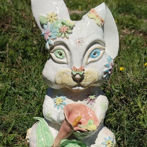 Op de afbeelding: Witte keramische konijnenfiguur versierd met kleurrijke bloemen en bladeren, die een roze bloem met een gele kern vasthoudt. Het konijn heeft blauwe en groene ogen en een vlinder op zijn neus.